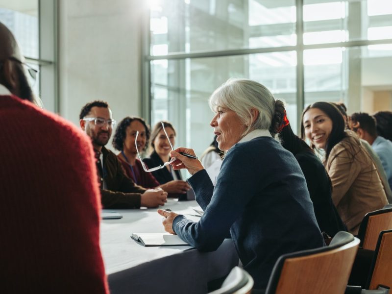 Diverse group of business professionals in a meeting with a woman speaking. Colleagues engage in a collaborative discussion in a modern conference room.