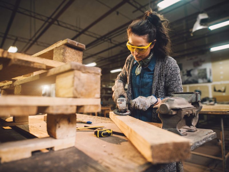 Portrait view of hardworking middle aged professional female carpenter worker working with sandpaper and choosing wood in the workshop.