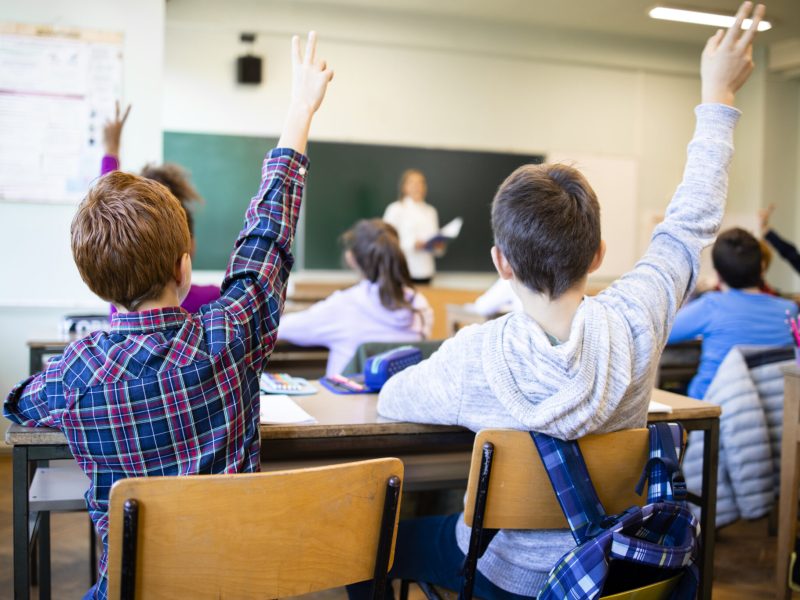 Schoolchildren at classroom with raised hands answering teacher's question.