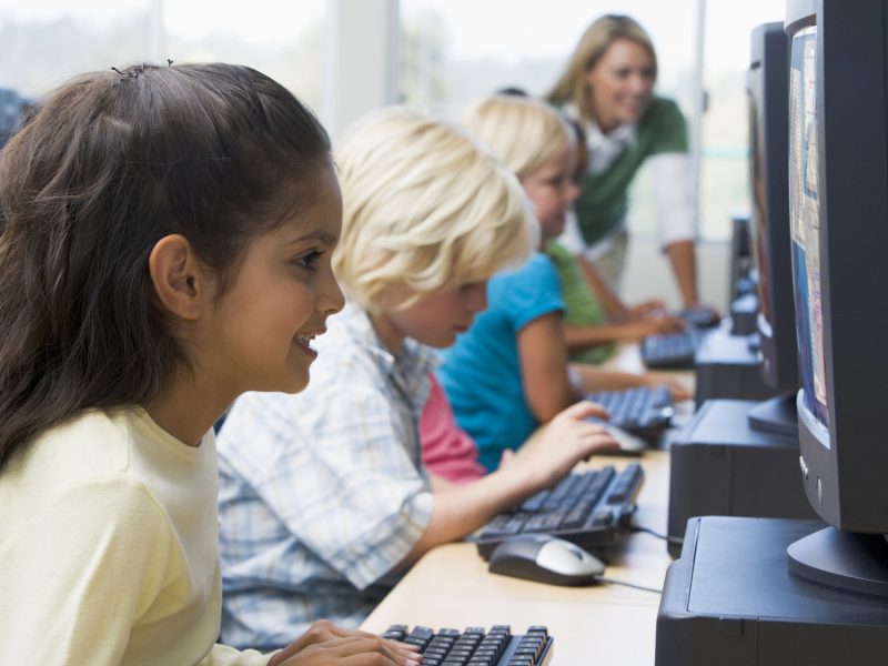 Children at computer terminals with teacher in background