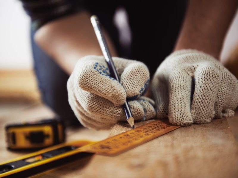 Close-up of craftsman hands in protective gloves measuring wooden plank with ruler and pencil. Woodwork and renovation concept.