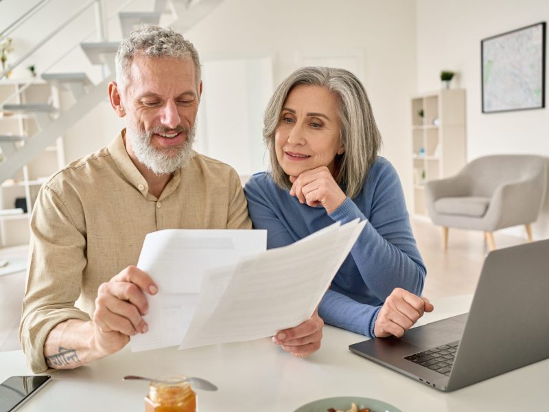 Middle aged couple reading paper bills calculating pension using laptop at home.