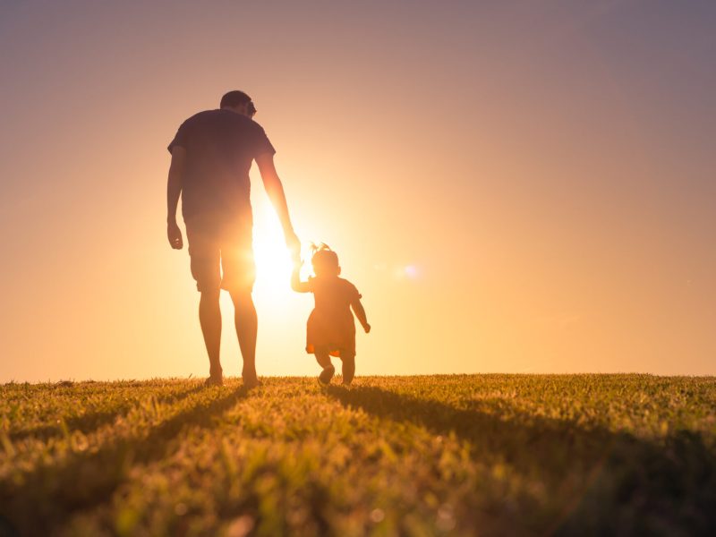 Father and daughter holding hands and walking at sunset outdoors.