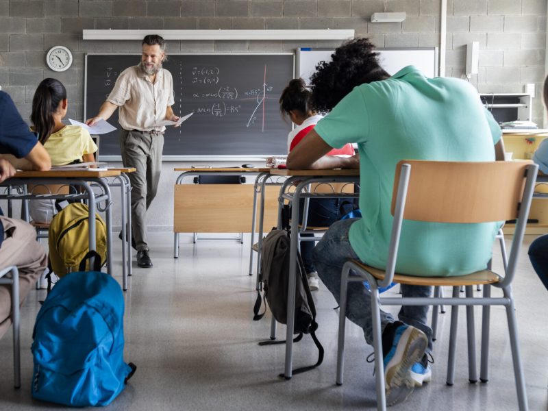 Mature caucasian man teacher hands out exams to multiracial high school students. Students ready to take exam.