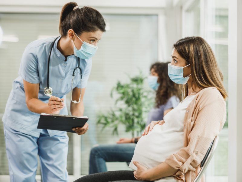 Pregnant Woman With Protective Mask Talking To Nurse At Waiting Room